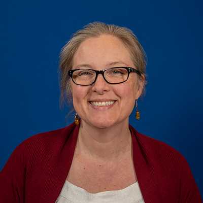 A middle-aged white woman with graying blonde hair, wearing glasses, a white blouse, and a dark red cardigan.