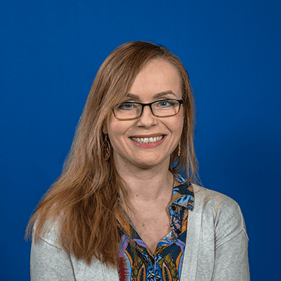 A white woman in her middle adulthood with mid-length blond hair and glasses, smiling at the camera.