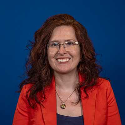 A white woman with auburn hair, wearing glasses and an orange blazer.