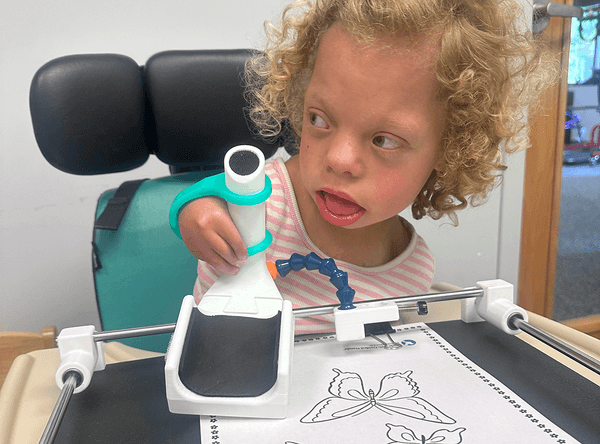 A young blonde girl with curly hair holds an assistive device that helps stabilize and guide hand movements. She is sitting in a chair with a tray. 