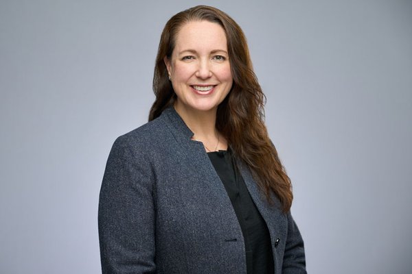 Smiling white woman with long brown hair. She is wearing a blue jacket and black top on a grey background.