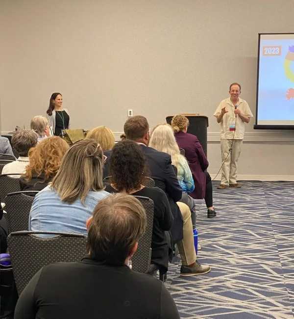 David Hoff speaks into the microphone to attendees while Jill Eastman (standing, far left) smiles and listens.