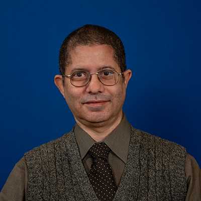 A middle-aged, Black male wearing eyeglasses, a gray sweater vest over a brown collated shirt, and tie with white dots.